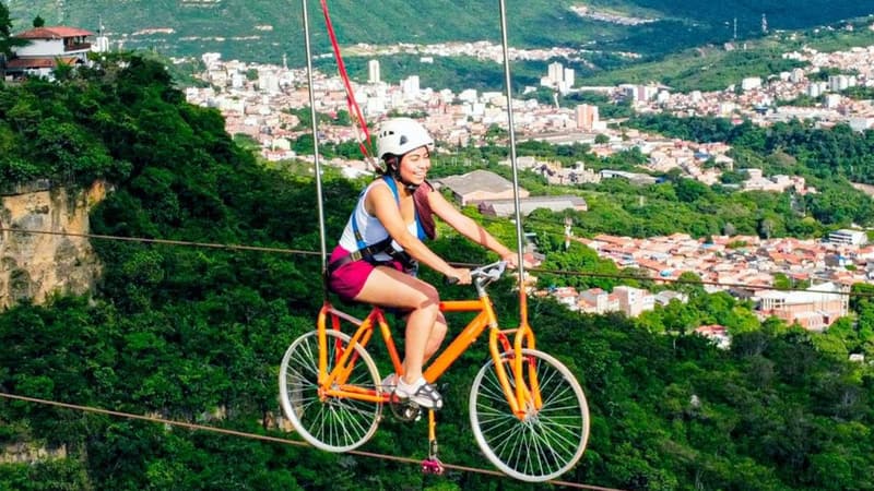Una ciclista pedaleando una bicicleta naranja sobre un cable alto, con San Gil al fondo.