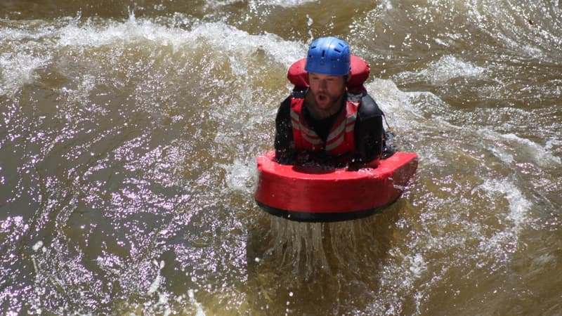 A hydrospeed rider in a helmet riding a red river board through a rapid.