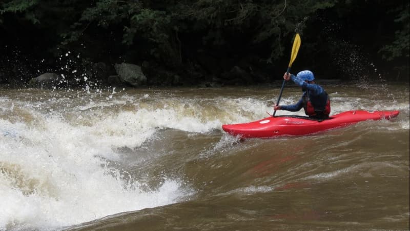A kayaker in a red boat surfing a whitewater wave.