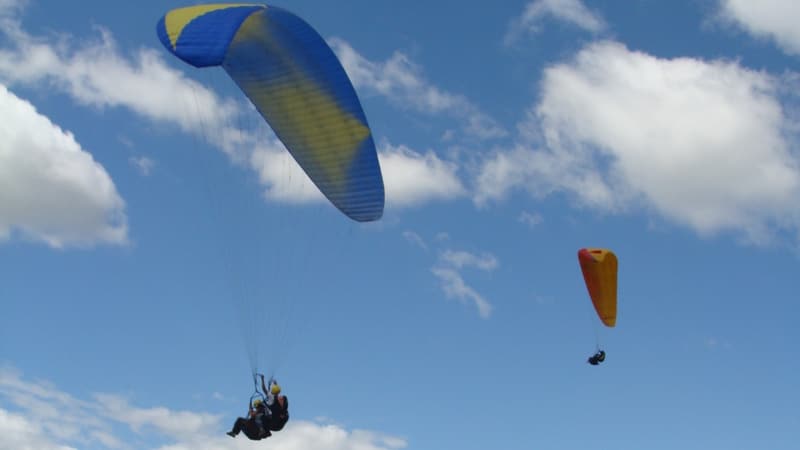 Dos parapentes flotando contra un cielo azul con nubes blancas.