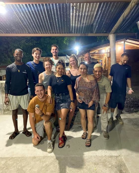 Eleven Macondo guests and friends grinning under a corrugated roof at a tejo club at night; one crouches at the front holding a tejo disc.