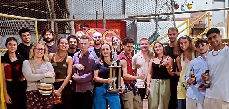A big group of travellers at a tejo club, one in the middle holding up a tall trophy.